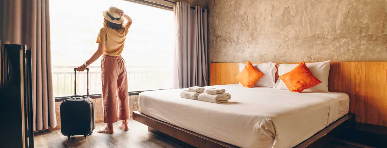 Woman looking out window of hotel next to bed with orange pillows